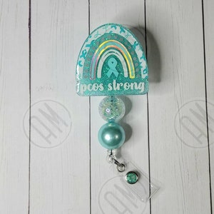 May include: Teal blue and white glitter resin rainbow with a teal blue ribbon and the text "pcos strong". The rainbow is attached to a teal blue and clear beaded retractable badge reel.