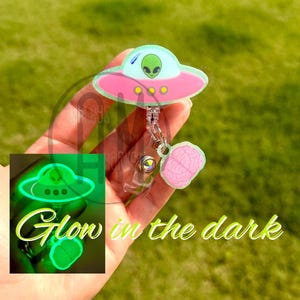 May include: A badge reel featuring a light green alien inside a pink and light green UFO, with a small pink brain charm attached. The UFO and brain glow in the dark, as shown in the inset image with the text Glow in the dark in green script.