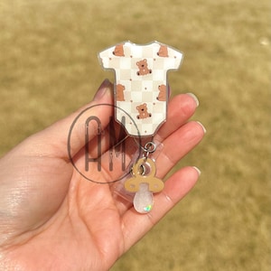 May include: A clear plastic badge holder with a brown teddy bear onesie design and a brown pacifier charm.