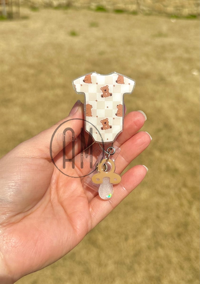 May include: A clear plastic badge holder with a brown teddy bear onesie design and a brown pacifier charm.