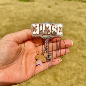 May include: A silver retractable badge holder with a clear plastic badge holder and a pacifier charm. The badge holder has a white background with the text "NURSE" in black letters and a pink and white pattern of baby faces.