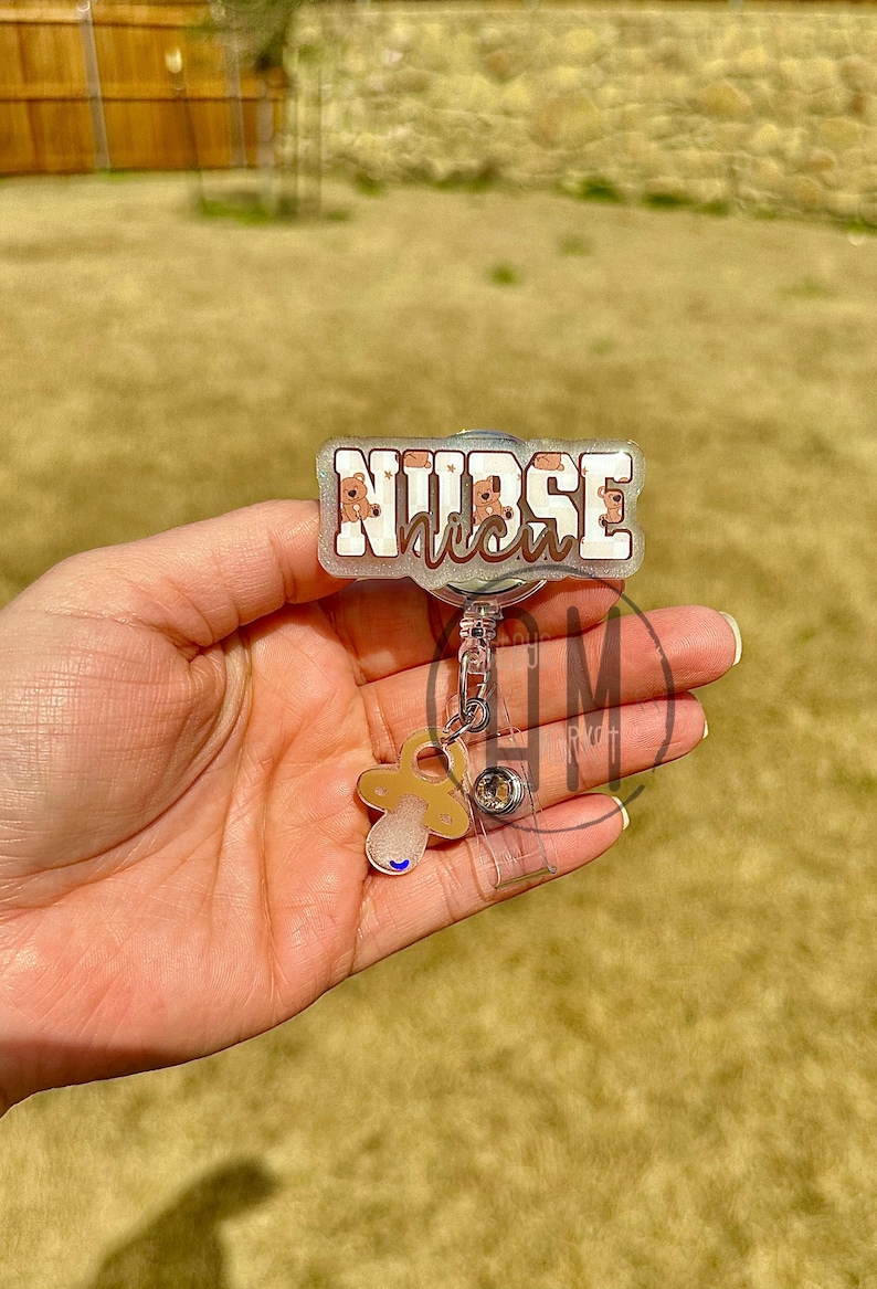 May include: A silver retractable badge holder with a clear plastic badge holder and a pacifier charm. The badge holder has a white background with the text "NURSE" in black letters and a pink and white pattern of baby faces.