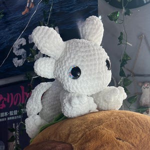 May include: A white crocheted axolotl plush toy with iridescent eyes sitting on top of a large brown plush toy.