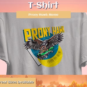 Op de afbeelding: Een grijs T-shirt met een grafische print van een skateboardende havik. De grafiek is in geel, blauw en rood. De tekst "PRONY HAWK" is in geel boven de grafiek gedrukt. De tekst "Prony Hawk Meme" is in geel gedrukt op een wit rechthoekje bovenaan het shirt.