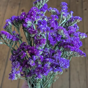 May include: A bunch of dried purple statice flowers. The flowers are a vibrant purple with small white accents, and the stems are a dried green colour. The flowers are bunched together, creating a full and textured appearance. The background is a wooden surface.