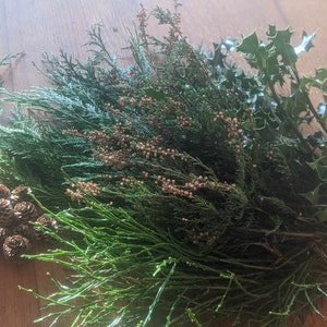 May include: A selection of fresh greenery, including holly, cedar, and small pine cones, presented on a wooden surface. The arrangement features various shades of green and brown, suitable for festive decorations or crafts.