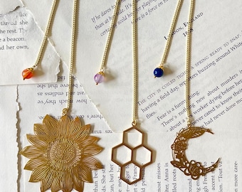 Könnte beinhalten: Vier goldene Lesezeichenketten mit verschiedenen Anhängern: eine Sonnenblume, eine Wabe, ein Halbmond und ein Blatt. Jede Kette hat einen kleinen Edelstein am Ende: orange, lila, blau und weiß.
