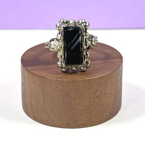May include: A silver-toned ring featuring a rectangular black stone set in a decorative metal frame with small bead-like accents along the edges. The setting is elongated and sits prominently on the band, showcasing a polished dark surface.