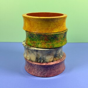 May include: Four decorative, cylindrical objects stacked on top of each other. The top object is yellow with a brown interior. Below it is a green and yellow object, followed by a marbled grey and tan object, and a reddish-brown object at the bottom.