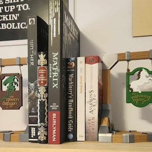 May include: A wooden bookend set with a medieval theme. The bookends feature a green dragon and a white prancing pony on signs hanging from the top. The bookends are holding up a variety of books, including "The Witcher", "The Matrix", "Assassin's Creed", and "Machinery's Handbook Guide".