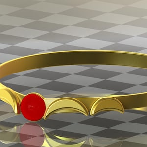 May include: A gold crown with a red jewel in the center. The crown is made of a thin metal band with a series of crescent shapes along the top.
