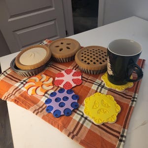 Peut inclure: Une collection de trois modèles de tartes miniatures dans des moules métalliques et quatre sous-verres circulaires aux motifs de fruits colorés, incluant orange, fraise, myrtille et citron, avec une tasse noire ornée d'un motif d'arbre blanc.