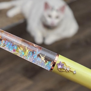 May include: A yellow pen with a clear barrel filled with glitter and a gold crown charm. The pen is being held by a hand.