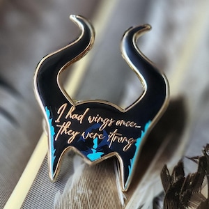 Puede incluir: Un pin de esmalte con borde dorado que presenta un dise&ntilde;o negro estilizado con cuernos curvos y detalles turquesa. El pin tiene la cita: "I had wings once... They were strong." El pin est&aacute; sobre un fondo de plumas.