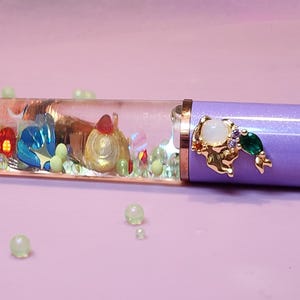 May include: A lavender fountain pen with a clear barrel filled with miniature colorful figures and a gold-colored tip. The pen has a gold and jewel accent near the nib and is set against a pink background with scattered beads.