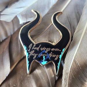Puede incluir: Un pin de esmalte negro y dorado con la silueta de cuernos y el texto "I had wings once... They were strong." El pin tiene detalles turquesa y azul, sobre un fondo de plumas marrones.
