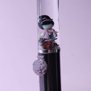 Puede incluir: Un bolígrafo transparente con una tapa negra y un encanto en forma de flor morada. Dentro del bolígrafo hay una pequeña figura de una niña que lleva un suéter rosa y un sombrero negro. También hay pequeñas piezas de brillo plateado y dorado dentro del bolígrafo.