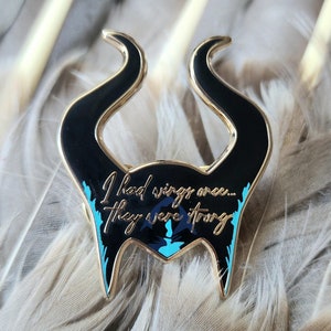 Puede incluir: Un pin de esmalte negro y dorado con forma de cuernos, con la cita "I had wings once... they were strong" en escritura dorada. El pin tiene detalles turquesa y est&aacute; sobre un fondo de plumas grises.