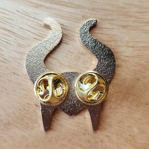 Puede incluir: Un pin de esmalte dorado con forma de cuernos estilizados. El pin tiene una superficie texturizada y dos cierres dorados. Los cuernos est&aacute;n curvados y apuntan hacia arriba. El pin es probablemente un accesorio decorativo.