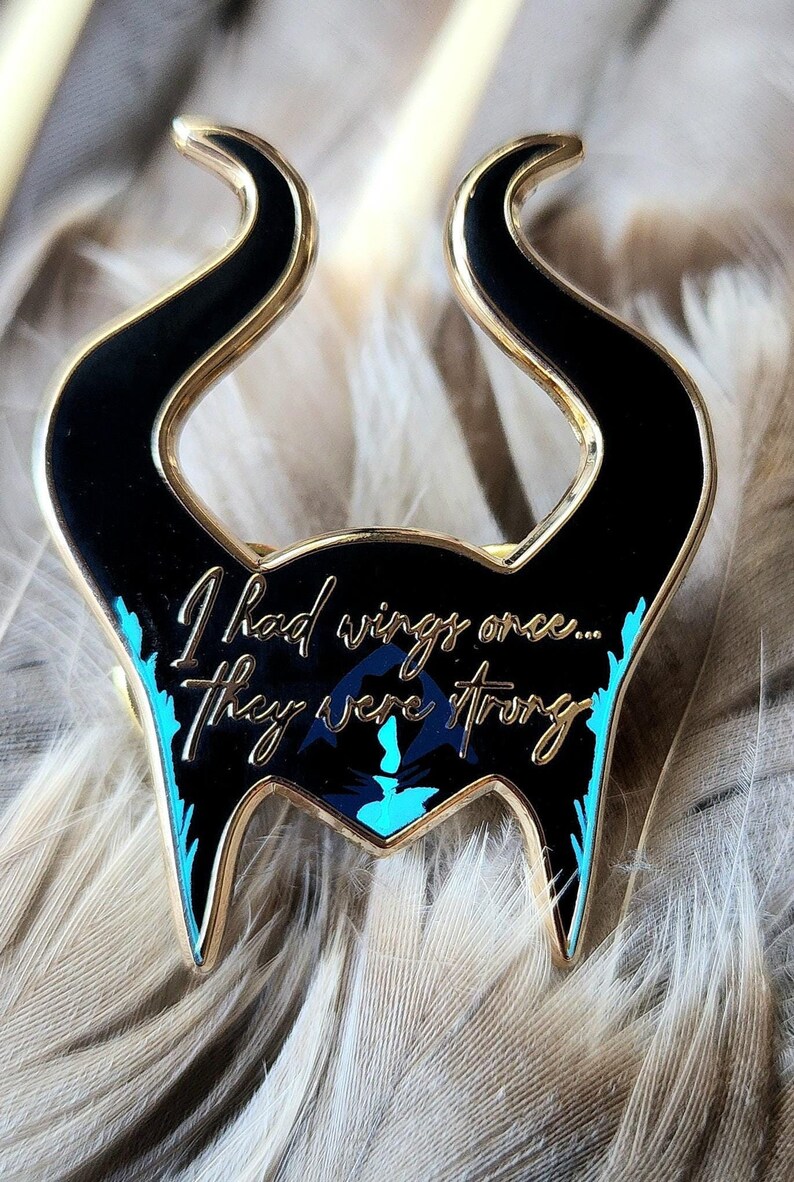 Puede incluir: Un pin de esmalte negro y dorado con forma de cuernos, con el texto "I had wings once... They were wrong." El pin tiene detalles turquesa y un dise&ntilde;o de silueta.