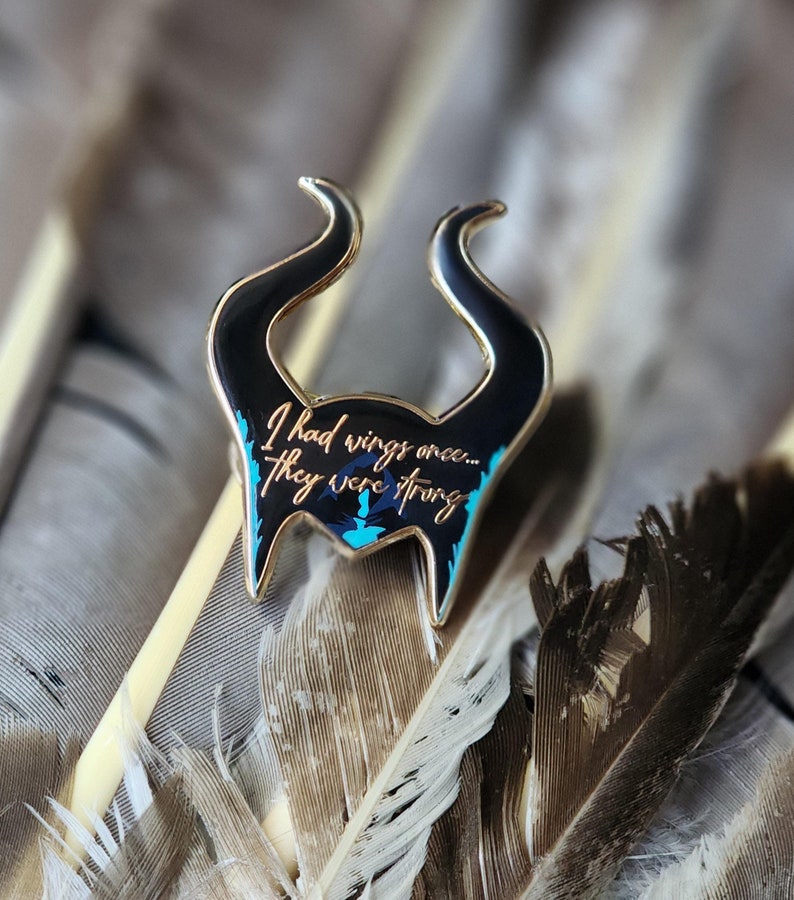Puede incluir: Un pin de esmalte con borde dorado que presenta un dise&ntilde;o negro estilizado con cuernos curvos y detalles turquesa. El pin tiene la cita: "I had wings once... They were strong." El pin est&aacute; sobre un fondo de plumas.
