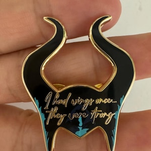 Puede incluir: Pin de esmalte negro y dorado con forma de cuernos estilizados. El pin presenta la cita "I had wings once... they were strong" en una elegante escritura con acentos turquesa. El pin tiene un borde dorado.