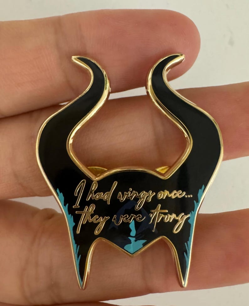 Puede incluir: Pin de esmalte negro y dorado con forma de cuernos estilizados. El pin presenta la cita "I had wings once... they were strong" en una elegante escritura con acentos turquesa. El pin tiene un borde dorado.
