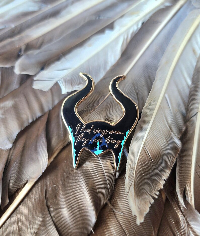 Puede incluir: Un pin de esmalte negro y dorado con la silueta de cuernos y el texto "I had wings once... They were strong." El pin tiene detalles turquesa y azul, sobre un fondo de plumas marrones.
