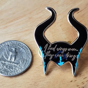 Puede incluir: Un pin de esmalte negro y dorado con forma de cuernos, con la cita "I had wings once... they were strong". El pin tiene detalles en azul y est&aacute; junto a una moneda de veinticinco centavos estadounidense.