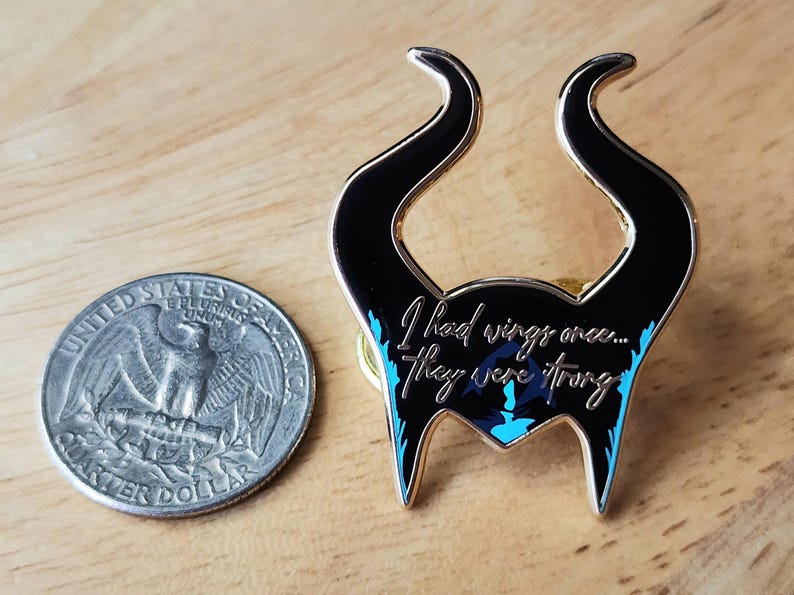 Puede incluir: Un pin de esmalte negro y dorado con forma de cuernos, con la cita "I had wings once... they were strong". El pin tiene detalles en azul y est&aacute; junto a una moneda de veinticinco centavos estadounidense.