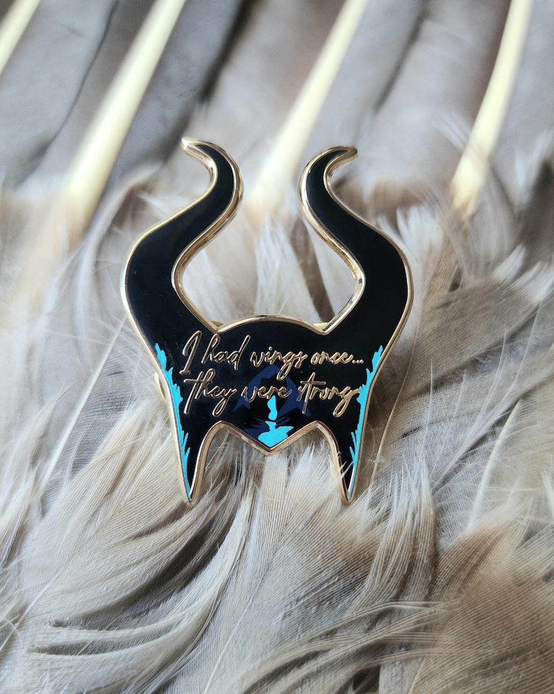 Puede incluir: Un pin de esmalte negro y dorado con forma de cuernos, con la cita "I had wings once... they were strong" en escritura dorada. El pin tiene detalles turquesa y est&aacute; sobre un fondo de plumas grises.