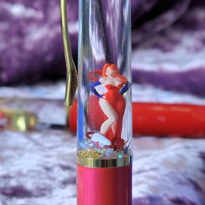 May include: A clear pen with a gold top and a red bottom. Inside the pen is a small figurine of a woman in a red dress. The pen is filled with glitter.