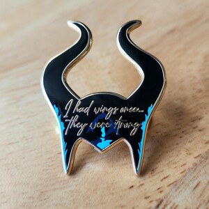 Puede incluir: Un pin de esmalte negro y dorado con forma de cuernos, con el texto "I had wings once... they were strong". El pin tiene detalles en azul y un borde dorado. El pin est&aacute; sobre una superficie de madera.