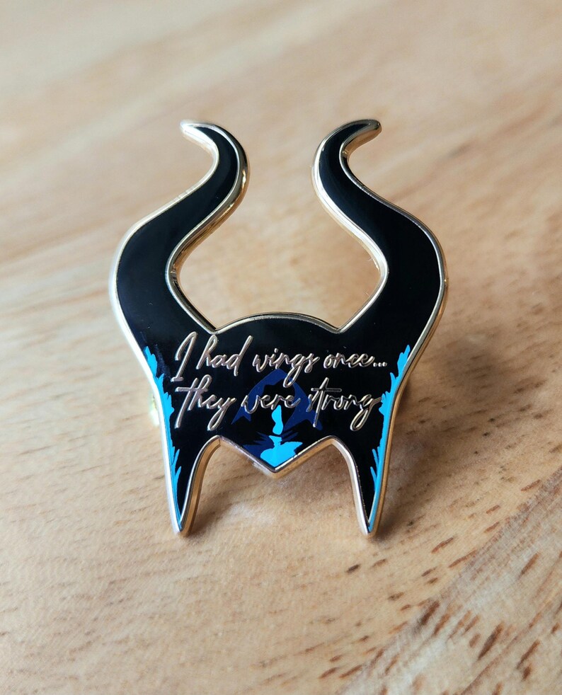 Puede incluir: Un pin de esmalte negro y dorado con forma de cuernos, con el texto "I had wings once... they were strong". El pin tiene detalles en azul y un borde dorado. El pin est&aacute; sobre una superficie de madera.