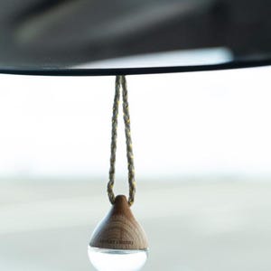 May include: A car air freshener hanging from a rearview mirror. The freshener has a wooden top and a clear, rounded bottom. The hanging cord is gray with gold accents. The text "DESERT EMBERS" is visible on the freshener.