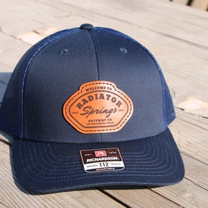 Może przedstawiać: Czapka trucker w kolorze granatowym z brązową skórzaną naszywką z napisem "Welcome to Radiator Springs, Since 1902, Gateway to the Ornament Valley". Czapka jest marki Richardson 112.