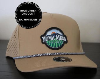 May include: A tan baseball cap with a perforated side panel and a rope detail across the brim. A circular patch features a landscape illustration with mountains, a sun, and green fields, with the text YUMA MESA FARMS. A black sticker is on the brim.