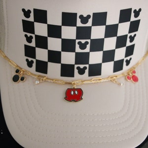 Puede incluir: Una gorra tipo trucker de color beige con un patr&oacute;n de cuadros blancos y negros y peque&ntilde;as siluetas de cabezas de rat&oacute;n negras. Una cadena dorada sujeta en la parte frontal incluye peque&ntilde;os dijes, como un pantal&oacute;n rojo y negro y orejas.
