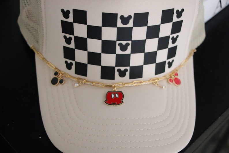 Puede incluir: Una gorra tipo trucker de color beige con un patr&oacute;n de cuadros blancos y negros y peque&ntilde;as siluetas de cabezas de rat&oacute;n negras. Una cadena dorada sujeta en la parte frontal incluye peque&ntilde;os dijes, como un pantal&oacute;n rojo y negro y orejas.