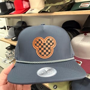May include: A navy blue baseball cap with a Mickey Mouse-shaped patch featuring a checkered design in orange and black. The cap has a white and grey rope detail along the brim and a curved peak. Other caps are visible on shelves in the background.