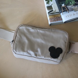 K&ouml;nnte beinhalten: Eine beige Bauchtasche mit einem schwarzen Mickey-Maus-Kopf-Patch und einem wei&szlig;en verstellbaren Gurt.