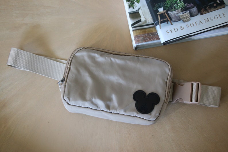 K&ouml;nnte beinhalten: Eine beige Bauchtasche mit einem schwarzen Mickey-Maus-Kopf-Patch und einem wei&szlig;en verstellbaren Gurt.