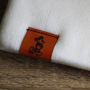 May include: White beanie with a brown leather patch featuring a black silhouette of Mickey Mouse.