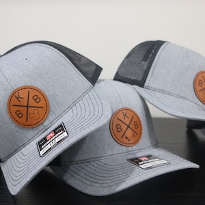 May include: Three grey and black baseball caps with brown leather patches. The patches have the letters KBB and a cross design.