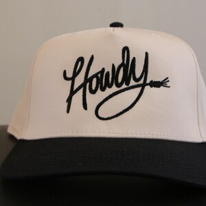 May include: A white baseball cap with a black embroidered design that says "Howdy" with a lasso. The cap has a black brim.