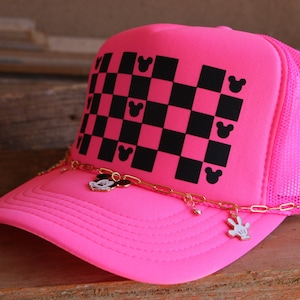 Puede incluir: Una gorra de camionero rosa ne&oacute;n con un patr&oacute;n de cuadros negros y rosas en el panel frontal, decorado con peque&ntilde;as siluetas de cabezas de rat&oacute;n negras. Una cadena dorada cuelga sobre la visera, adornada con peque&ntilde;os dijes met&aacute;licos de rat&oacute;n.