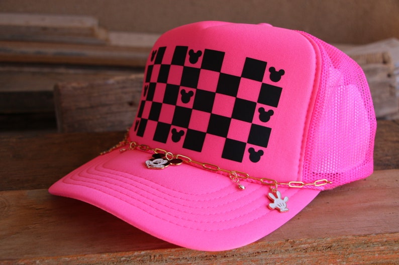 Puede incluir: Una gorra de camionero rosa ne&oacute;n con un patr&oacute;n de cuadros negros y rosas en el panel frontal, decorado con peque&ntilde;as siluetas de cabezas de rat&oacute;n negras. Una cadena dorada cuelga sobre la visera, adornada con peque&ntilde;os dijes met&aacute;licos de rat&oacute;n.