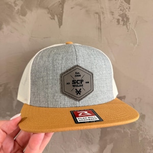 Op de afbeelding: Een snapback pet met een grijze kroon, bruine klep en witte mesh achterkant. De pet heeft een leren patch met de tekst "SCP WELDZ" en "Est. 2008". De klep is effen bruin van kleur.