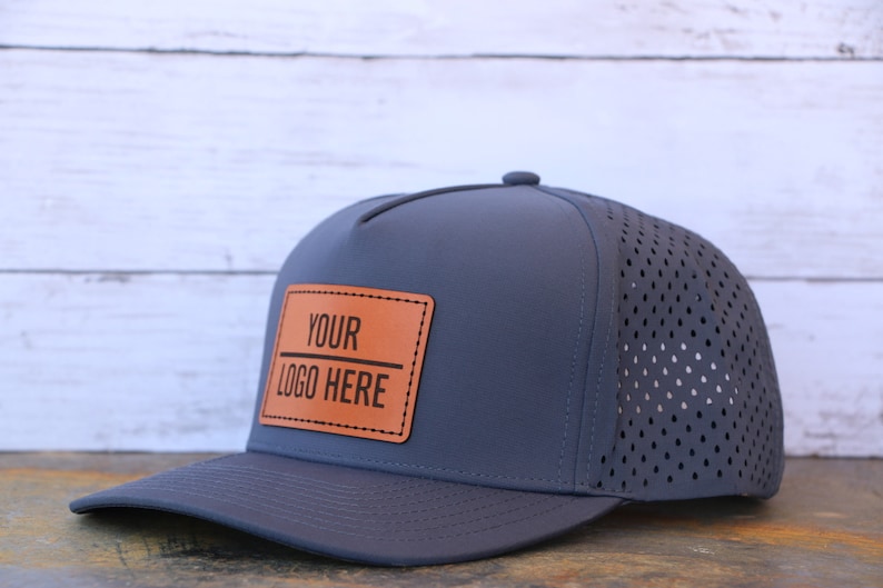 May include: A dark gray baseball cap featuring a rectangular tan leather patch on the front panel. The patch is stitched with black text that reads "YOUR LOGO HERE" with a horizontal line in between. The cap has a perforated back panel.