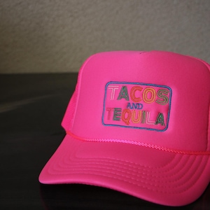 May include: Pink trucker hat with a black mesh back. The hat has a colorful embroidered patch that says "Tacos and Tequila".